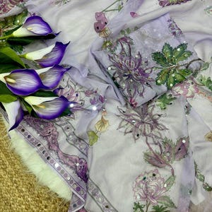 May include: A light purple fabric with floral patterns in shades of purple, green, and white. The fabric has a decorative border with mirrored accents. Purple calla lilies are arranged on the left side of the image.