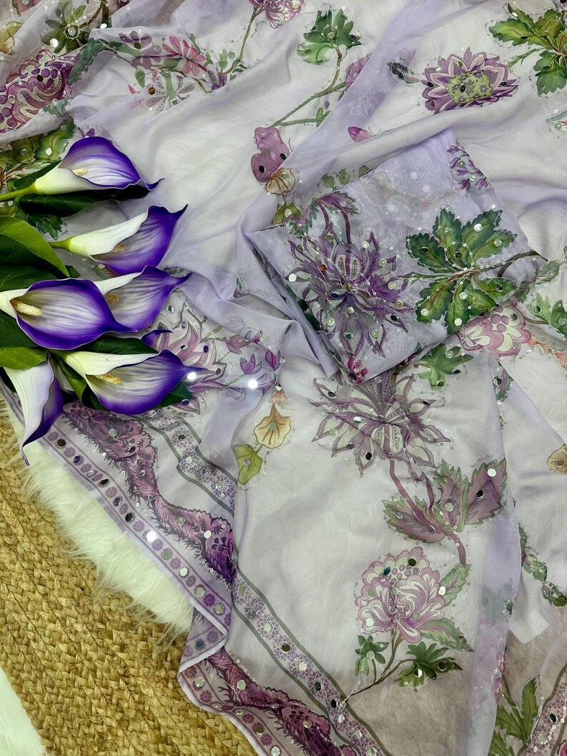May include: A light purple fabric with floral patterns in shades of purple, green, and white. The fabric has a decorative border with mirrored accents. Purple calla lilies are arranged on the left side of the image.