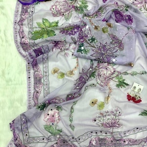 May include: A sheer, lavender-colored fabric with a floral pattern in shades of purple, pink, and green. The fabric is adorned with small, reflective embellishments along the edges and on the floral designs. A matching piece of fabric is folded nearby.