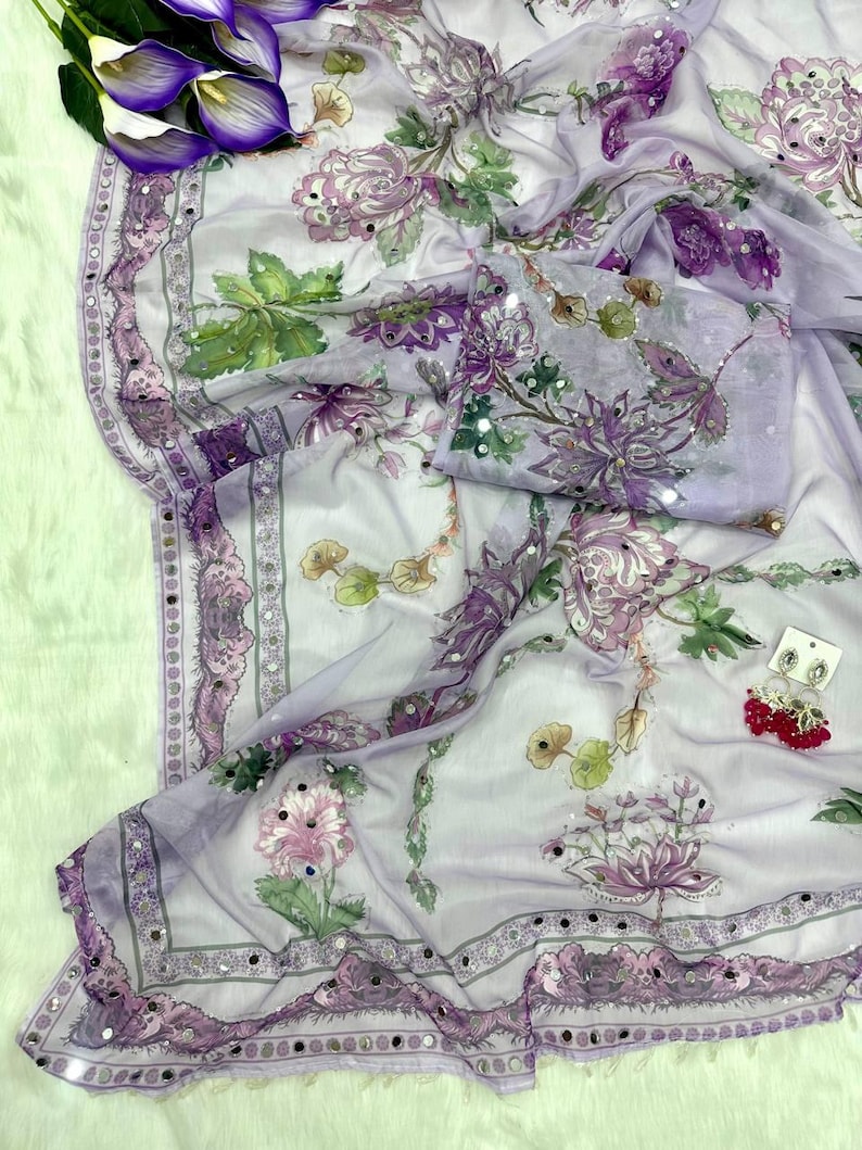 May include: A sheer, lavender-colored fabric with a floral pattern in shades of purple, pink, and green. The fabric is adorned with small, reflective embellishments along the edges and on the floral designs. A matching piece of fabric is folded nearby.