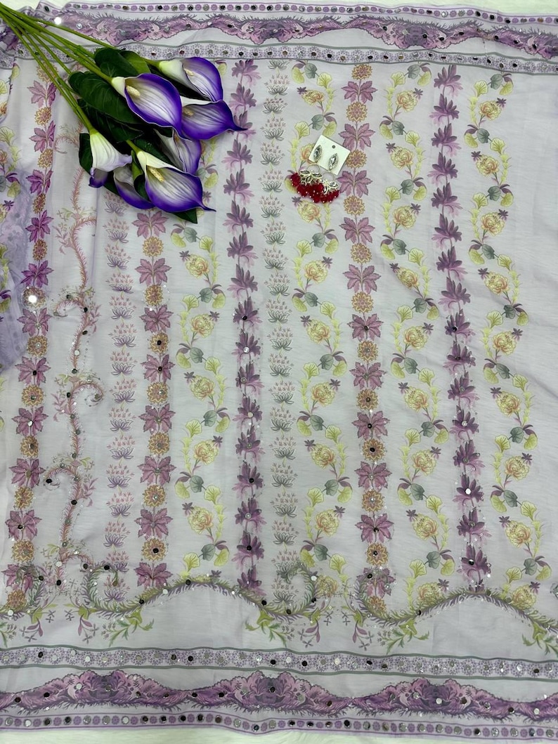 May include: A light purple fabric with a floral pattern in shades of purple, yellow, and green. The fabric has a decorative border with mirrored accents. A bouquet of purple calla lilies and a pair of earrings are placed on the fabric.