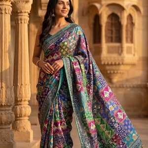 May include: A colorful, patterned saree with a teal border and intricate embroidery. The saree features a patchwork design with shades of blue, green, purple, and pink. The fabric has a shimmering quality, and the border is embellished with small mirrors.