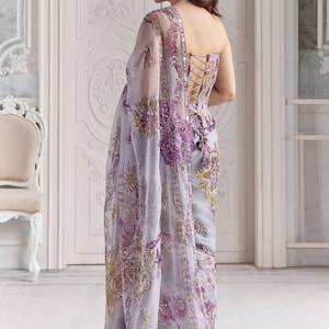 May include: A light purple saree with intricate floral embroidery in shades of purple, gold, and green. The saree features a corset-style bodice with lace-up detailing and a sheer, embellished shawl. The fabric is adorned with sequins.