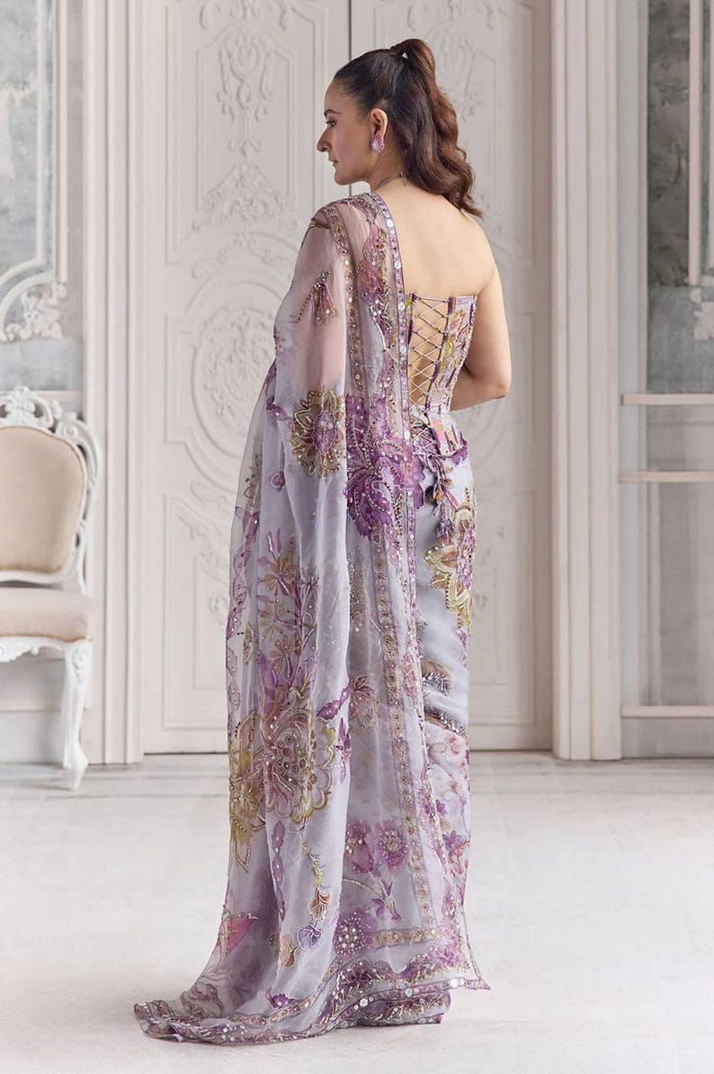 May include: A light purple saree with intricate floral embroidery in shades of purple, gold, and green. The saree features a corset-style bodice with lace-up detailing and a sheer, embellished shawl. The fabric is adorned with sequins.