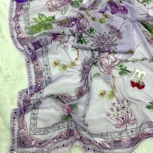 May include: A light purple saree with a floral pattern in shades of purple, green, and white. The saree has a decorative border with small mirrored accents. The fabric is draped, and a matching blouse piece is included.