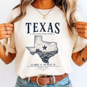 Peut inclure: Un t-shirt couleur crème avec le texte TEXAS THE LONE STAR STATE en bleu foncé. En dessous se trouve une carte du Texas contenant une illustration de paysage et une étoile. Le texte 31.9686 N, 99.9018 W, TEXAS, USA, EST. 1845 est imprimé.
