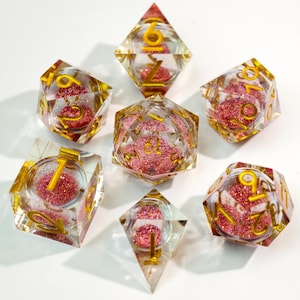 Pode incluir: Um conjunto de sete dados poliédricos feitos de resina transparente, cada um contendo um núcleo de glitter rosa. Os dados apresentam números de cor dourada gravados nas superfícies. O conjunto inclui várias formas, incluindo d20, d12, d10, d8, d6 e d4.