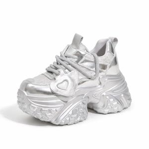 May include: Silver chunky sneaker with a heart-shaped detail on the side. The shoe features a thick, textured sole and a padded tongue. The laces are thick and silver, matching the rest of the shoe.