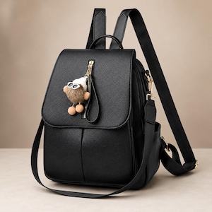 Könnte beinhalten: Schwarzer Rucksack aus Kunstleder mit Klappenverschluss und Reißverschlusstasche vorne. Der Rucksack hat verstellbare Schultergurte und einen abnehmbaren Crossbody-Riemen. Ein kleiner, flauschiger Anhänger mit Sonnenbrille hängt am Reißverschluss.