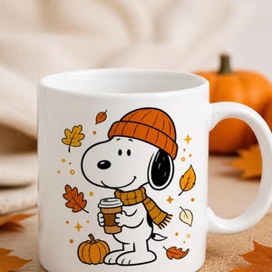 May include: A white ceramic mug featuring a graphic illustration of Snoopy wearing an orange knit beanie and a matching scarf. The character holds a coffee cup, surrounded by orange and yellow autumn leaves, small stars, and a pumpkin.