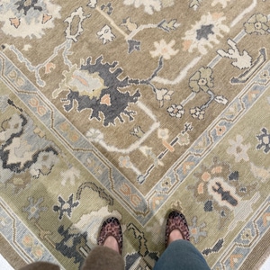 Puede incluir: Una alfombra grande con una base de color verde oliva apagado y un intrincado patrón de flores y enredaderas en tonos gris carbón, crema suave y melocotón pálido. El diseño incluye un borde decorativo ancho con motivos geométricos.