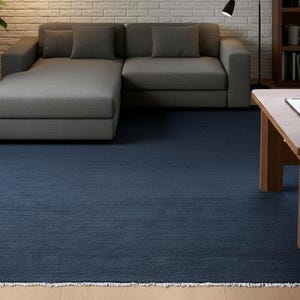 May include: A large, rectangular navy blue area rug with a fringed edge. The rug covers most of the floor in a living room setting, with a gray sectional sofa, wooden coffee table, and a bookshelf visible in the background.