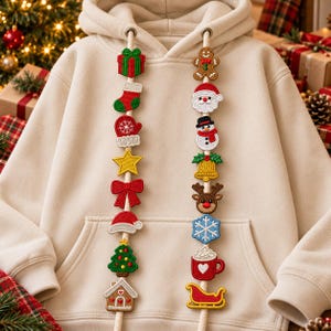 May include: A beige hooded sweatshirt featuring two white drawstrings decorated with embroidered Christmas-themed patches, including a gift, stocking, mitten, star, bow, Santa hat, tree, gingerbread house, gingerbread man, Santa, snowman, bell, reindeer, snowflake, mug, and sleigh.