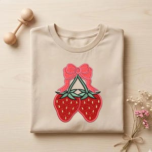 May include: A folded, beige t-shirt featuring a design of two red strawberries with green leaves and a pink bow. The strawberries have a glittery texture. A wooden rattle and a small bouquet of flowers are also visible.