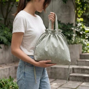 May include: A sage green drawstring pouch bag featuring a subtle white floral pattern. The bag has two top handles and a gathered, ruffled opening secured by thin matching fabric cords that hang down the sides of the soft, rounded structure.