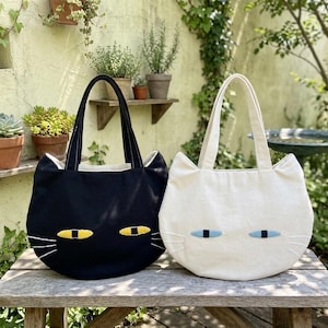 Puede incluir: Dos bolsos tote con forma de cabeza de gato, uno negro y otro blanco roto. El bolso negro presenta ojos amarillos y bigotes bordados en blanco, mientras que el bolso blanco tiene ojos azul claro y bigotes bordados en blanco.