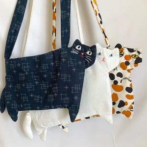 以下が含まれることがあります： 猫の形をした3つの布製ショルダーバッグ。1つは白の格子柄が入ったダークブルー、1つは無地の白、もう1つは黒、オレンジ、白の斑点がある三毛猫柄です。それぞれのバッグには刺繍で顔のパーツが施されています。