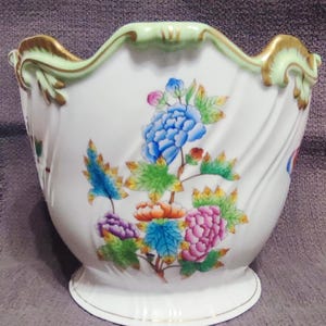 Pode incluir: Um vaso de porcelana branca com borda recortada em verde claro e detalhes dourados. O corpo é decorado com um padrão floral vibrante, incluindo flores azuis, rosas, laranjas e roxas com folhas verdes sobre uma superfície lisa e curva.