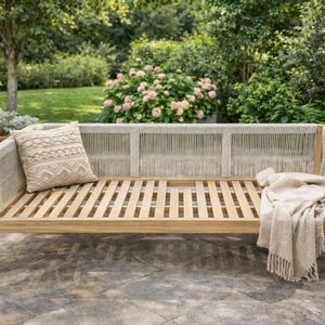 Può includere: Un divano da esterno in legno con schienale intrecciato e seduta a doghe. Una coperta beige e un cuscino decorativo sono appoggiati sul divano. Il divano è circondato da verde e fiori.