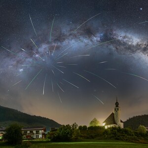 Peut inclure: Paysage nocturne avec une église sous un ciel étoilé et la Voie lactée. De nombreuses traînées lumineuses, probablement des météores, rayonnent d'un point central. Le paysage comprend un village et des collines verdoyantes.