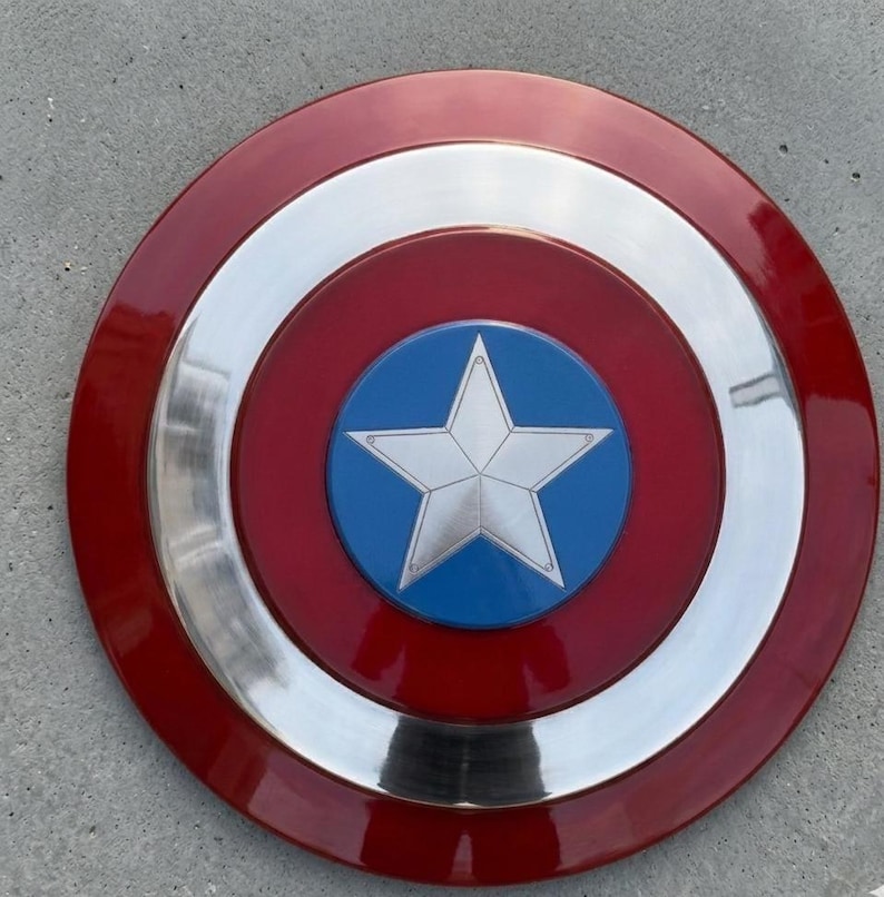 May include: A circular shield in red, white, and blue. The design includes a silver star on a blue circle at the centre, encircled by rings of red, silver, and red. This shield is a replica.