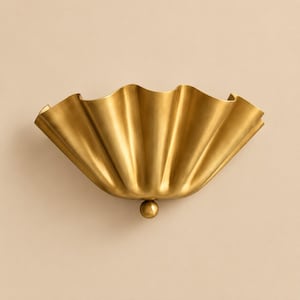 May include: A semi-circular wall sconce featuring a metallic gold finish with a pleated, fan-like design. The fixture has a wide, undulating top edge that flares outward and a small, spherical knob detail at the bottom center point.
