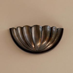May include: A bronze-colored, wall-mounted decorative sconce with a scalloped, shell-like design. The sconce has a semi-circular shape with multiple vertical, curved ridges, creating a textured surface. The dark bronze finish adds a touch of elegance.