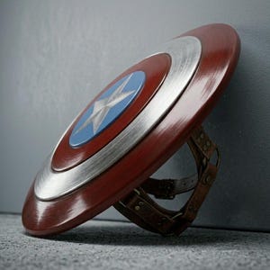 May include: A red, white, and blue shield with a star emblem, a classic superhero accessory. The shield features a metallic silver ring and brown leather straps on the back.