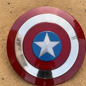 May include: A circular shield featuring a red outer ring, a silver band, and a blue central disc with a white star. The shield has a shiny surface and is likely a replica.