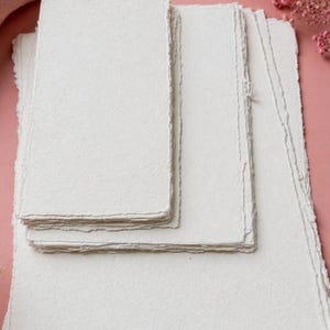 May include: A collection of off-white handmade paper sheets with uneven, deckled edges. The paper is stacked in different sizes, showing a textured surface. The background is a soft pink, with a few sprigs of pink flowers in the top right corner.