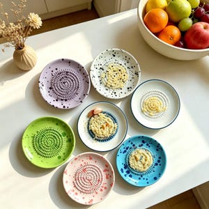 Peut inclure: Une collection de râpes à ail en céramique colorées. Les râpes sont de différentes couleurs, notamment violet, vert, rose et bleu, avec une surface de râpage en spirale. Certaines râpes ont des mouchetures noires. Un bol de fruits est en arrière-plan.