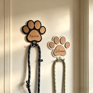 May include: Two wooden paw print-shaped leash holders with names Raina and Silvana. The paw prints have a light brown centre and a black and white border. Each holder has a metal hook and a matching leash. The Raina leash is navy blue, and the Silvana leash is green.