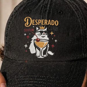 May include: A black washed-denim baseball cap featuring a graphic of a white cat wearing a gold crown and black sunglasses. The cat holds a cocktail glass with a cherry. The text reads Desperado Club and So Fun, surrounded by small star icons.
