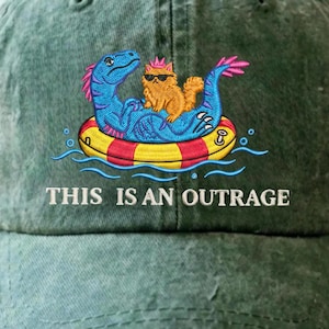May include: A forest green baseball cap featuring an embroidered graphic of a blue dinosaur wearing sunglasses and a pink crown, carrying an orange cat on a red and yellow striped pool float, with the text THIS IS AN OUTRAGE printed below.