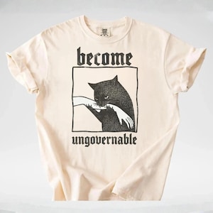 May include: Cream-colored t-shirt featuring a black graphic of a cat biting a hand, framed by a rectangle. The words "become" and "ungovernable" are printed above and below the graphic in a gothic font. The shirt has short sleeves.