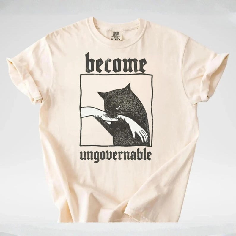 May include: Cream-colored t-shirt featuring a black graphic of a cat biting a hand, framed by a rectangle. The words "become" and "ungovernable" are printed above and below the graphic in a gothic font. The shirt has short sleeves.