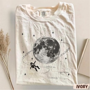 Könnte beinhalten: Ein elfenbeinfarbenes T-Shirt mit einer schwarzen Grafik, die einen großen Mond und einen kleinen Astronauten im Weltraum zeigt. Der Text lautet: I'M AN ASTRONAUT, I STARE AT YOU, I SING TO YOU, I CIRCLE YOU, YOU'RE THE MOON. Kleine Sterne sind auf dem Stoff verteilt.
