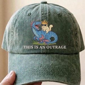 May include: A washed olive green baseball cap featuring an embroidered graphic of a blue dinosaur with a small tan cat wearing a gold crown and black sunglasses on its back, with the text THIS IS AN OUTRAGE printed in white capital letters.