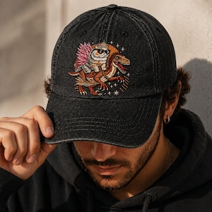 May include: Black denim baseball cap with an embroidered design of a cat in sunglasses riding a dinosaur. The cat has a pink feathered headdress. The dinosaur is orange and brown. The cap has a curved peak and a button on top.