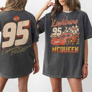 May include: A charcoal gray t-shirt featuring a graphic of the red race car Lightning McQueen with the number 95, a checkered flag, and the text Lightning McQueen. The back of the shirt displays a large number 95 and the Rust-eze logo.