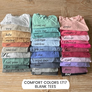 May include: Stacks of blank Comfort Colors 1717 t-shirts in various colors, including white, blue spruce, and black. Each stack is labeled with the color name in a handwritten font. The shirts are neatly folded and arranged on a wooden surface.