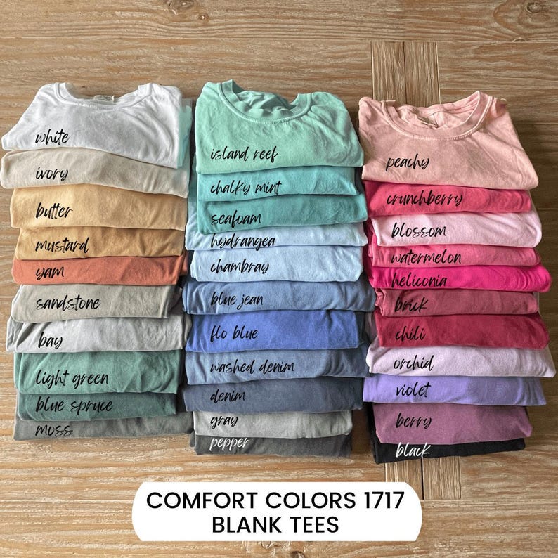 May include: Stacks of blank Comfort Colors 1717 t-shirts in various colors, including white, blue spruce, and black. Each stack is labeled with the color name in a handwritten font. The shirts are neatly folded and arranged on a wooden surface.