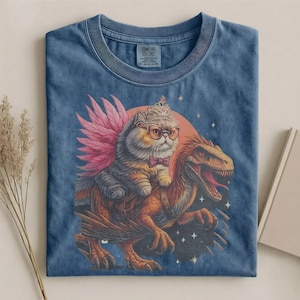 May include: Blue Comfort Colors t-shirt featuring a whimsical graphic of a cat wearing glasses, a crown, and a bow tie, riding a dinosaur with pink wings. The design is set against a red circle and starry background.