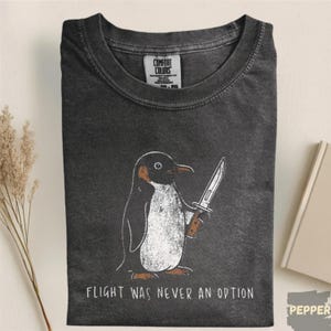May include: A charcoal grey crew neck t-shirt featuring a graphic of a penguin holding a knife. The text below the penguin reads, "FLIGHT WAS NEVER AN OPTION." The shirt has a Comfort Colors brand label on the inside of the collar area.