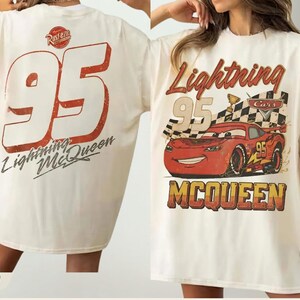 May include: An ivory t-shirt featuring a graphic of the red race car Lightning McQueen from the movie Cars. The front displays the car, a checkered flag, and the text Lightning McQueen. The back shows the number 95 and the text Lightning McQueen.