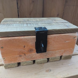 May include: A rectangular wooden box constructed from light-coloured planks with a hinged lid. The front panel features a prominent black metal latch mechanism centred on the exterior. The base is supported by small wooden blocks.