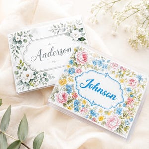 May include: Two personalized clear plastic wedding invitation holders. One features the name "Anderson" in script within a floral frame of white flowers and green leaves. The other has the name "Johnson" in blue script surrounded by pink, blue, and yellow flowers.