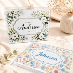 May include: Two personalized name card holders. One features the name "Anderson" in elegant script, framed by a floral design with white flowers and green foliage. The second holder displays "Johnson" within a floral border of pink, blue, and yellow flowers.
