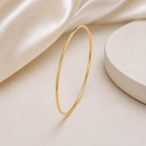 Könnte beinhalten: Ein zartes, goldfarbenes Armreif-Armband. Das dünne, kreisförmige Armband wird vor einem Hintergrund aus weichem, cremefarbenem Stoff und einer minimalistischen, weißen Plattform präsentiert. Das schlichte Design und die polierte Oberfläche des Armbands schaffen eine raffinierte Ästhetik.