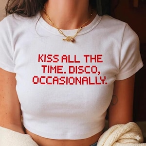 May include: White crop top featuring the red text "KISS ALL THE TIME. DISCO, OCCASIONALLY." A gold-coloured chain necklace is worn. The short-sleeved top has a round neckline.